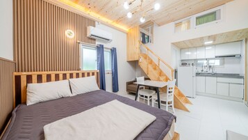 Un dormitorio con cama, mesita de noche, una silla, ventana con cortinas y una escalera que lleva a una zona de cocina.