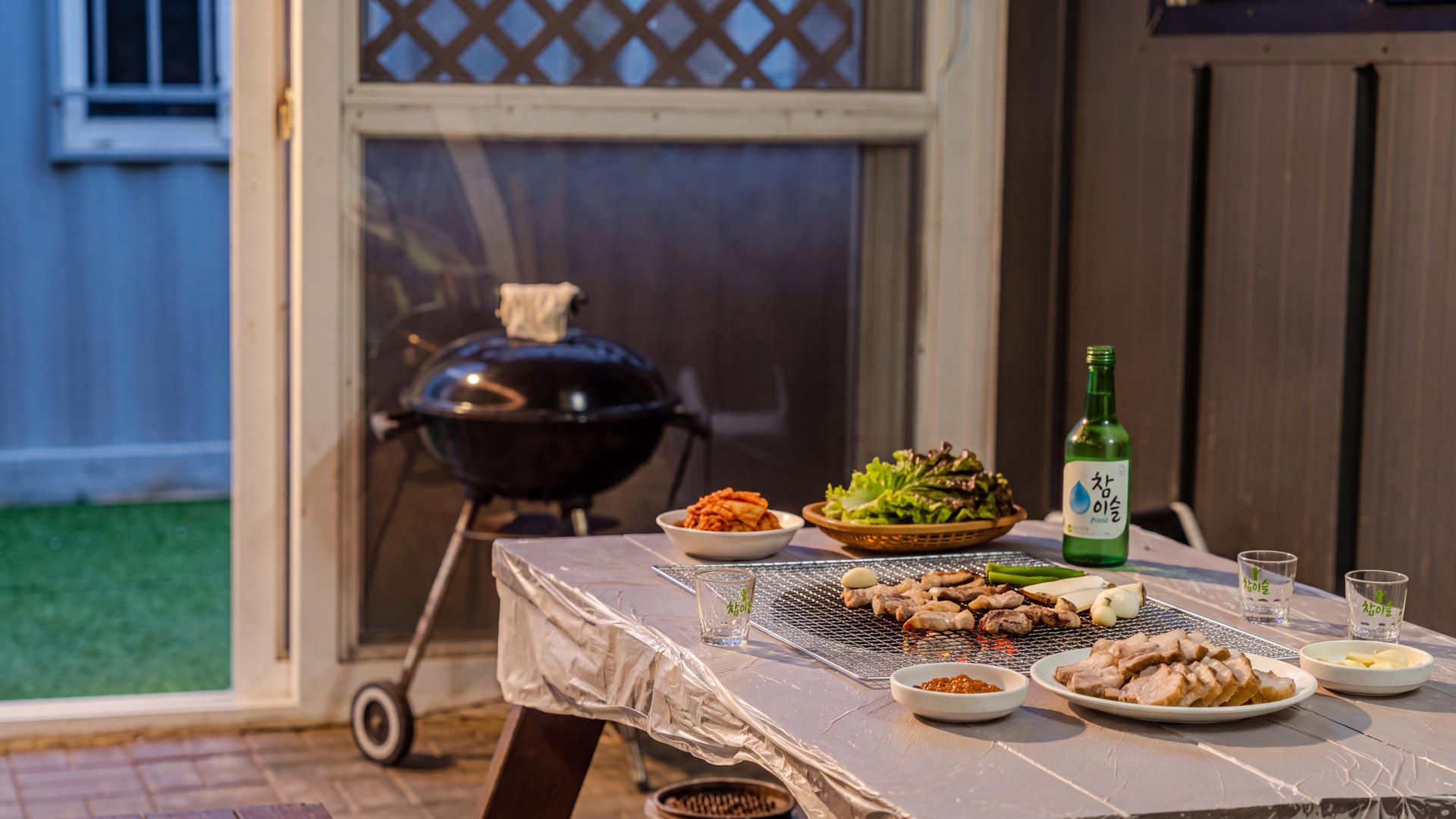 A barbecue setup with various meats, vegetables, and a bottle of green beverage on a covered table.