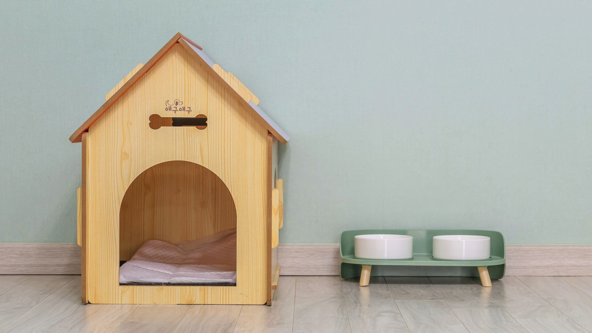 Una caseta de madera para perros con techo y entrada, junto a un comedero verde con dos cuencos.
