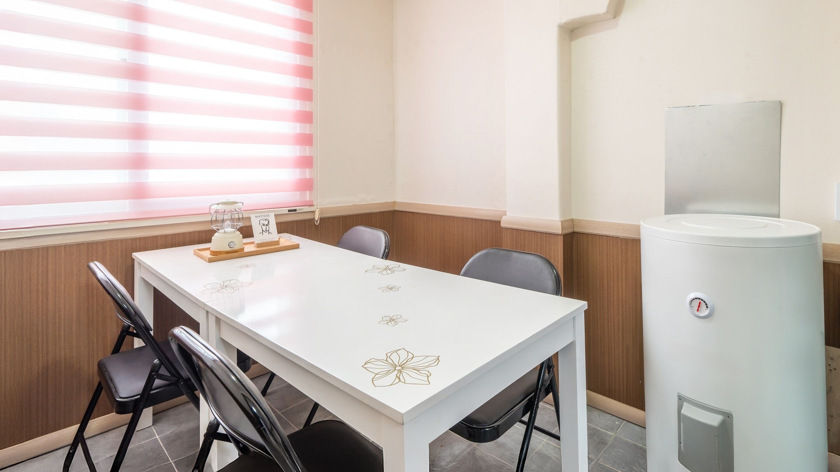 Una sala de reuniones pequeña con una mesa blanca, cuatro sillas, un dispensador de agua y un ventanal con persianas.