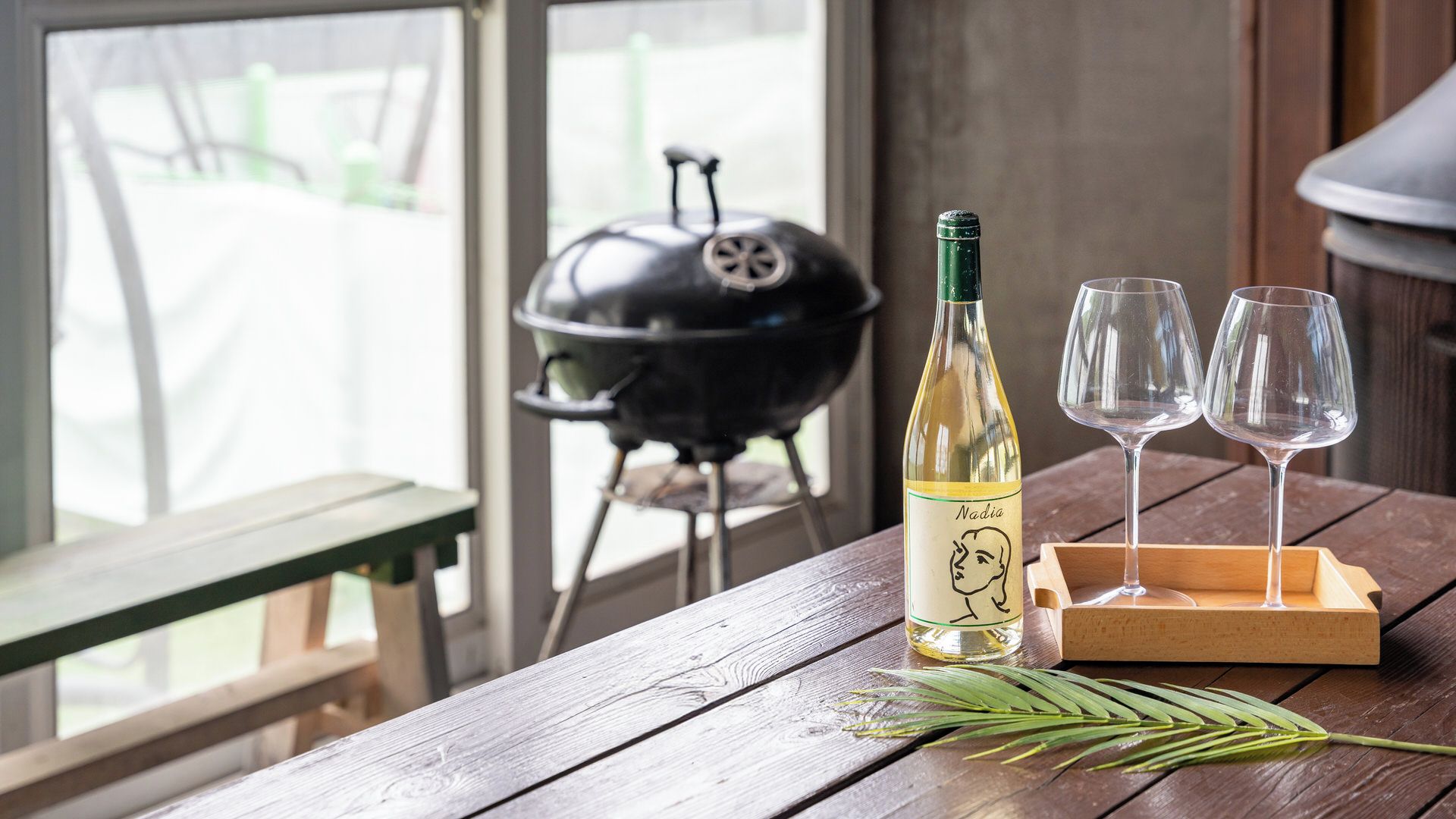 A wooden table with a bottle of white wine, two empty glasses, and a green plant. In the background, there is a barbecue grill.