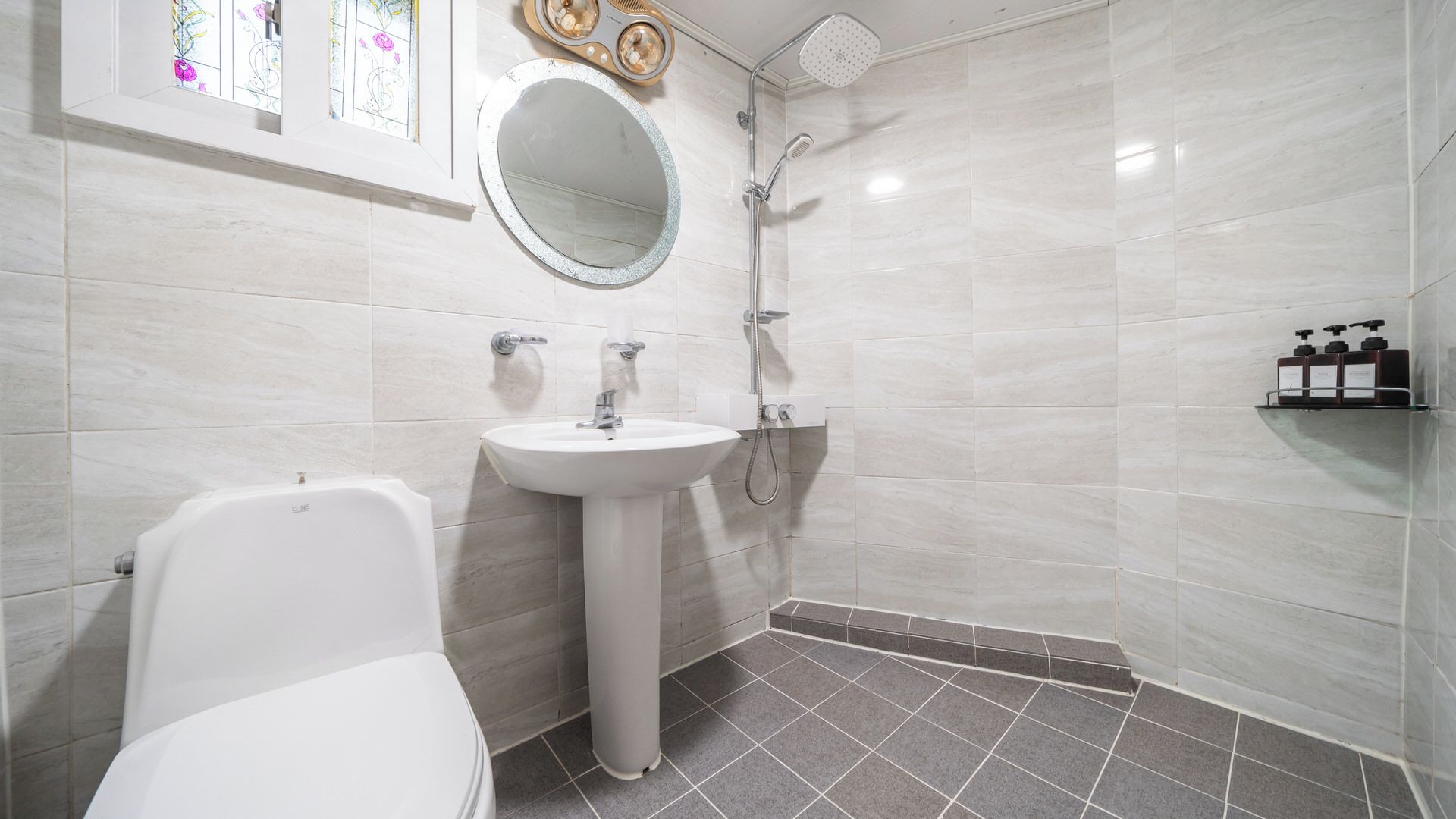 A modern bathroom with a white toilet, a pedestal sink, a shower with a handheld showerhead, and a mirror above the sink.