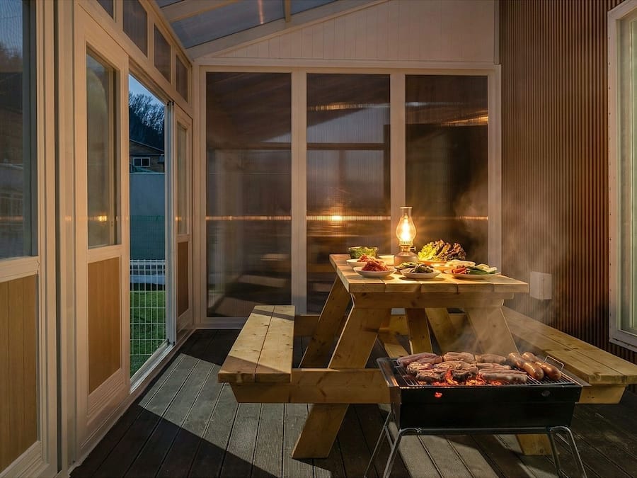 A wooden barbecue grill with food, a table with plates, and a lit lantern on a deck.