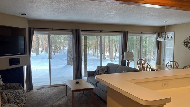 A cozy living room with a sofa, a small table, and a TV. There is a large window with curtains, a dining table with chairs, and a kitchen area with a counter. - Two Bedroom, Two Bath Condo ready for year-around fun. Great Up North Getaway! (Bellaire)