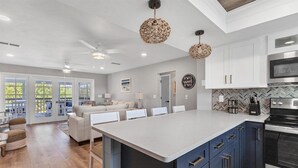 A modern kitchen with a central island, dark blue cabinets, and stainless steel appliances. There is a dining area with white chairs and a sofa.