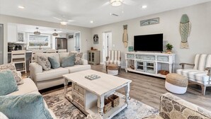 A spacious living room with a large sofa, a coffee table, and a television. There is a kitchen area visible in the background.