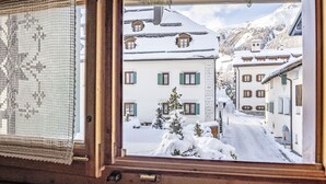 A window view of a snow-covered street with buildings, a wooden structure, and a mountain in the background.