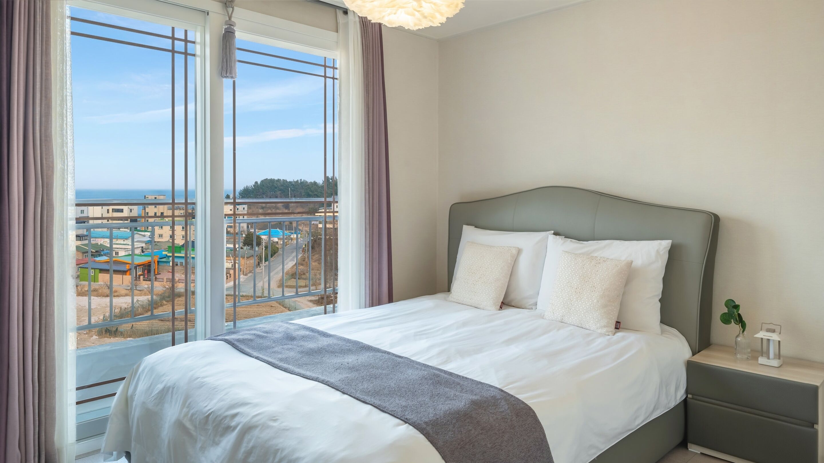 A hotel room with a bed, a nightstand, and a view of a cityscape through large windows.