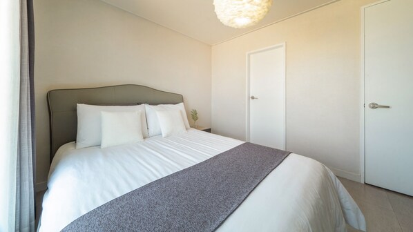 Una cama bien hecha con sábanas blancas y un plaid gris, una planta en la mesita de noche y una puerta blanca en una habitación sencilla y limpia.
