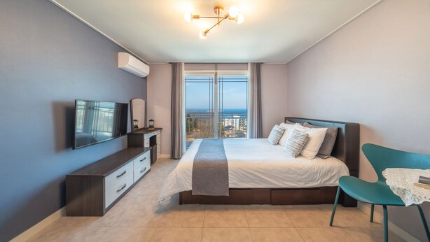 A modern bedroom with a large bed, a TV, a dresser, and a view of the city through large windows.