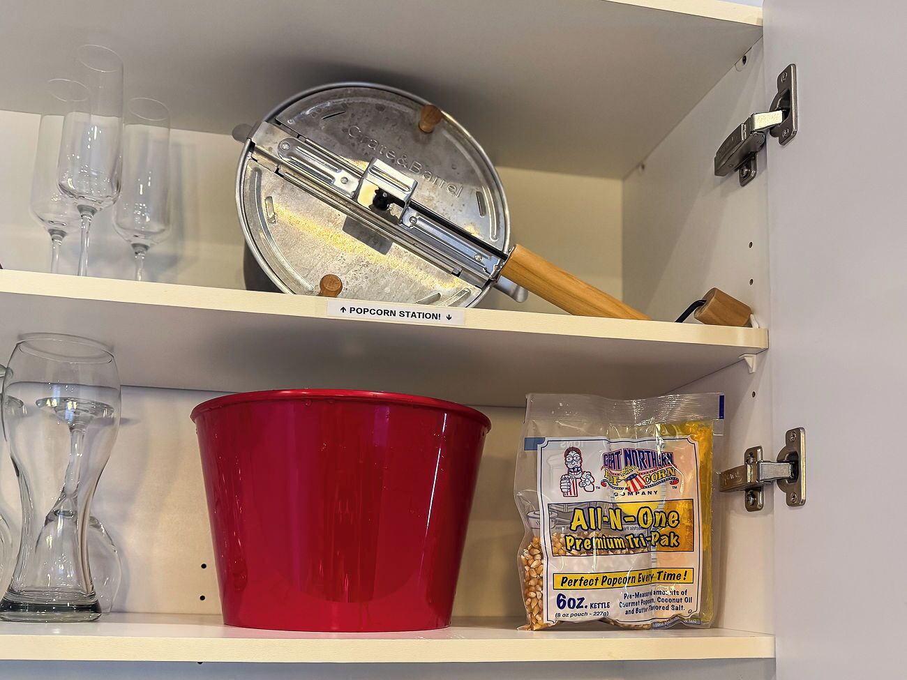 A kitchen shelf with a popcorn maker, a red bowl, a bag of popcorn seasoning, and three wine glasses.