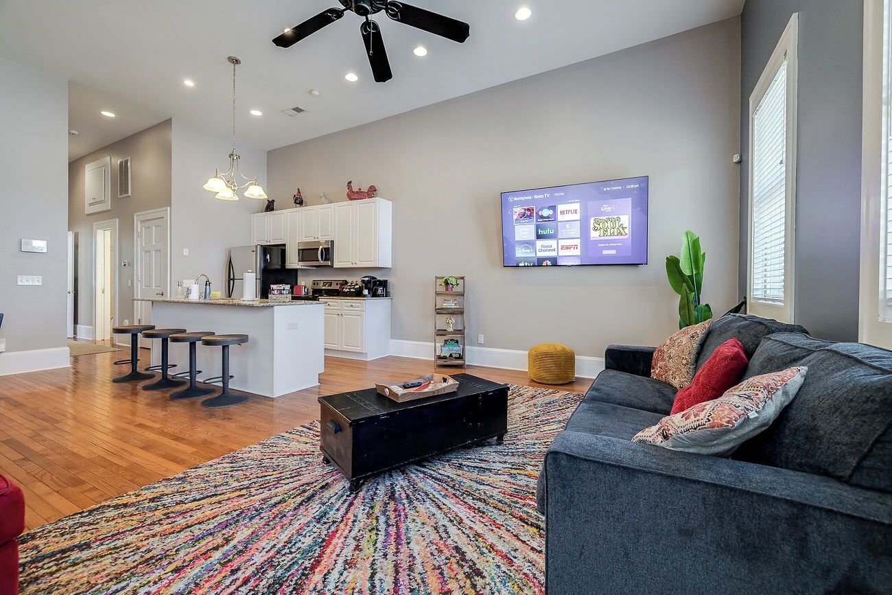 A modern living room with a gray sofa, colorful rug, and a black coffee table. A flat-screen TV is mounted on the wall, and a ceiling fan is visible.