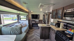 A compact RV interior with a sofa, kitchenette, TV, and a window with a view of a house and street. - Country Cowboy RV Campsite in River Ranch 72 (River Ranch)