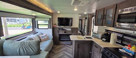 Interior compacto de una autocaravana con sofá, kitchenette, televisión y ventana con vista a una casa y una calle.