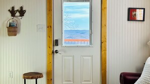 Una porta blanca amb una finestra, una cistella de vimet a la paret, un petit tamboret i un quadre amb marc.