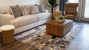 Een woonkamer met een bank, salontafel en fauteuil, een plafondventilator en een groot raam met jaloezieën.
