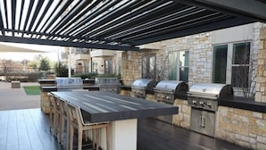 Een buitenkeuken met een lange tafel en meerdere barbecues, gelegen tegen een achtergrond van een stenen muur.