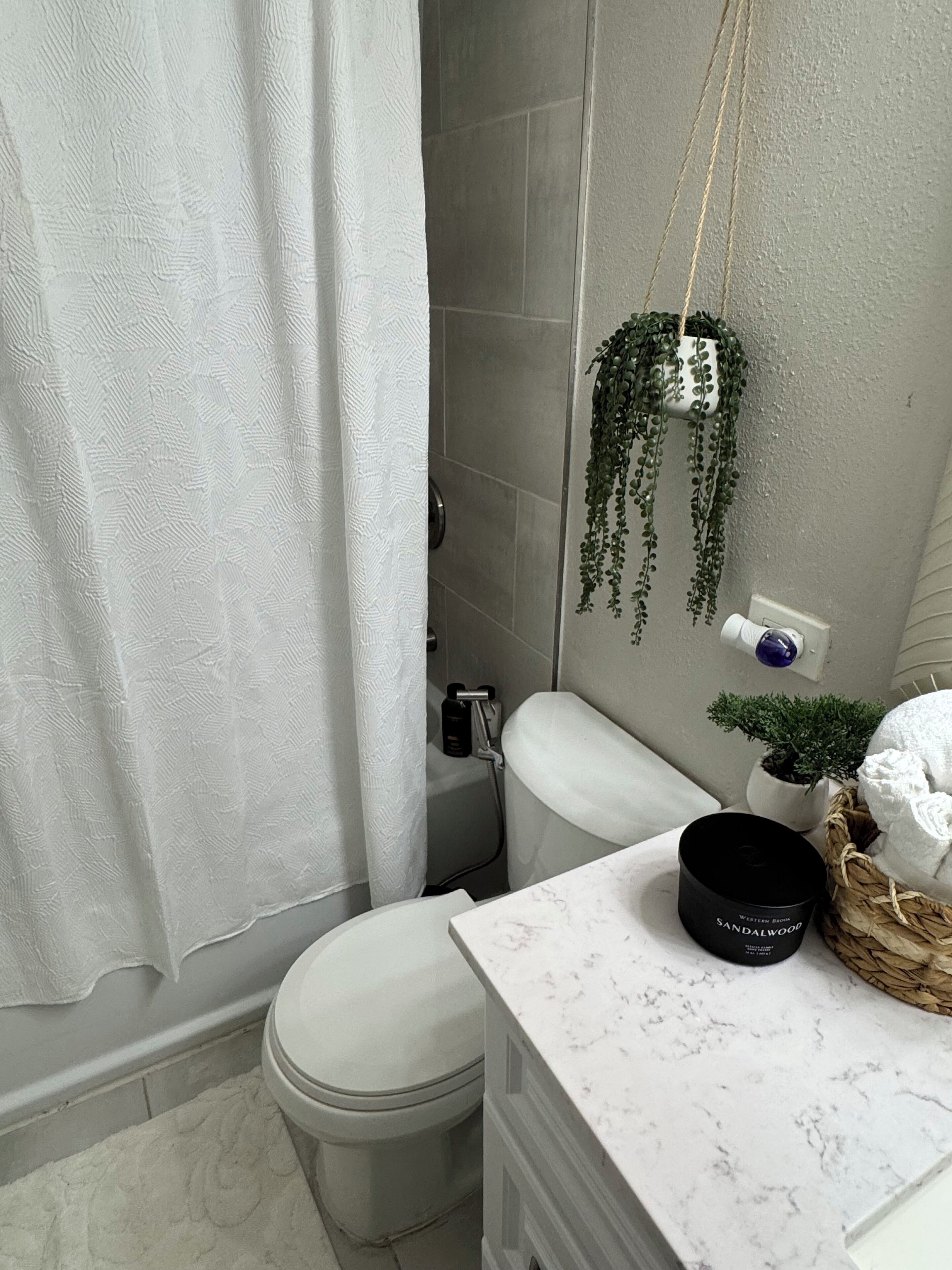 Ein Badezimmer mit Toilette, einer Duschvorhang, einer hängenden Pflanze, einem kleinen Topfpflanzen, einer Kerze und einem Korb mit Handtüchern.