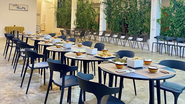 Een ruime eetruimte met tafels en stoelen, een hoog plafond met metalen balken en hanglampen.
