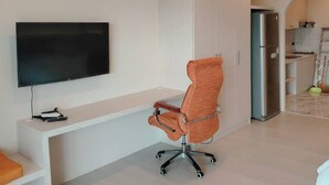 A modern living room with a flat-screen TV mounted on the wall, a white console table, an orange office chair, a built-in kitchen area with a refrigerator, and recessed ceiling lights. - Studio View Talay (Pattaya)