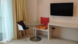 A modern, minimalist room with a round glass table, a single chair, a flat-screen TV mounted on the wall, and a window with sheer curtains. - Studio View Talay (Pattaya)