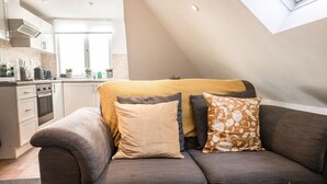 Een moderne woonkamer met een grijze bank, een gele plaid en een bloemenkussen. Op de achtergrond is een keuken zichtbaar met witte kastjes en een raam.