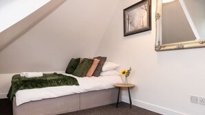 Een slaapkamer met een bed, kussens, een nachtkastje met een bloevas, een spiegel en een vloerbedekking.