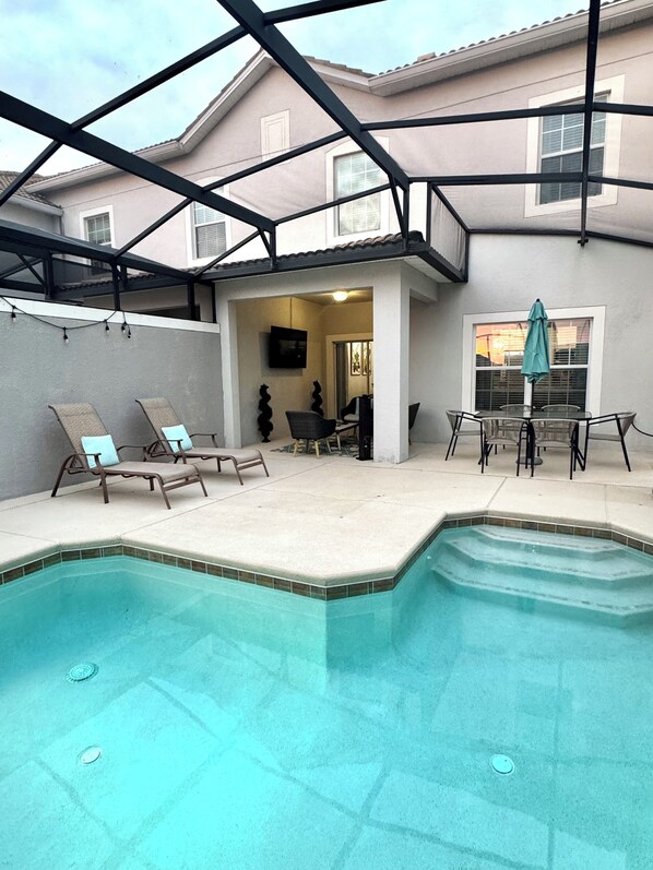 A covered outdoor swimming pool area with lounge chairs and a table. - Storey Lake Family DISNEY/UNIVERSAL EPIC UNIVERSE home w/ POOL and Lake View (Kissimmee)
