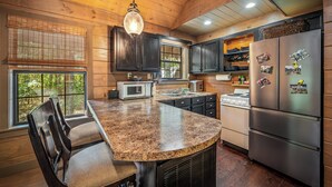 Una cocina con armarios de madera, encimera de granito, refrigerador de acero inoxidable y un comedor con una silla.