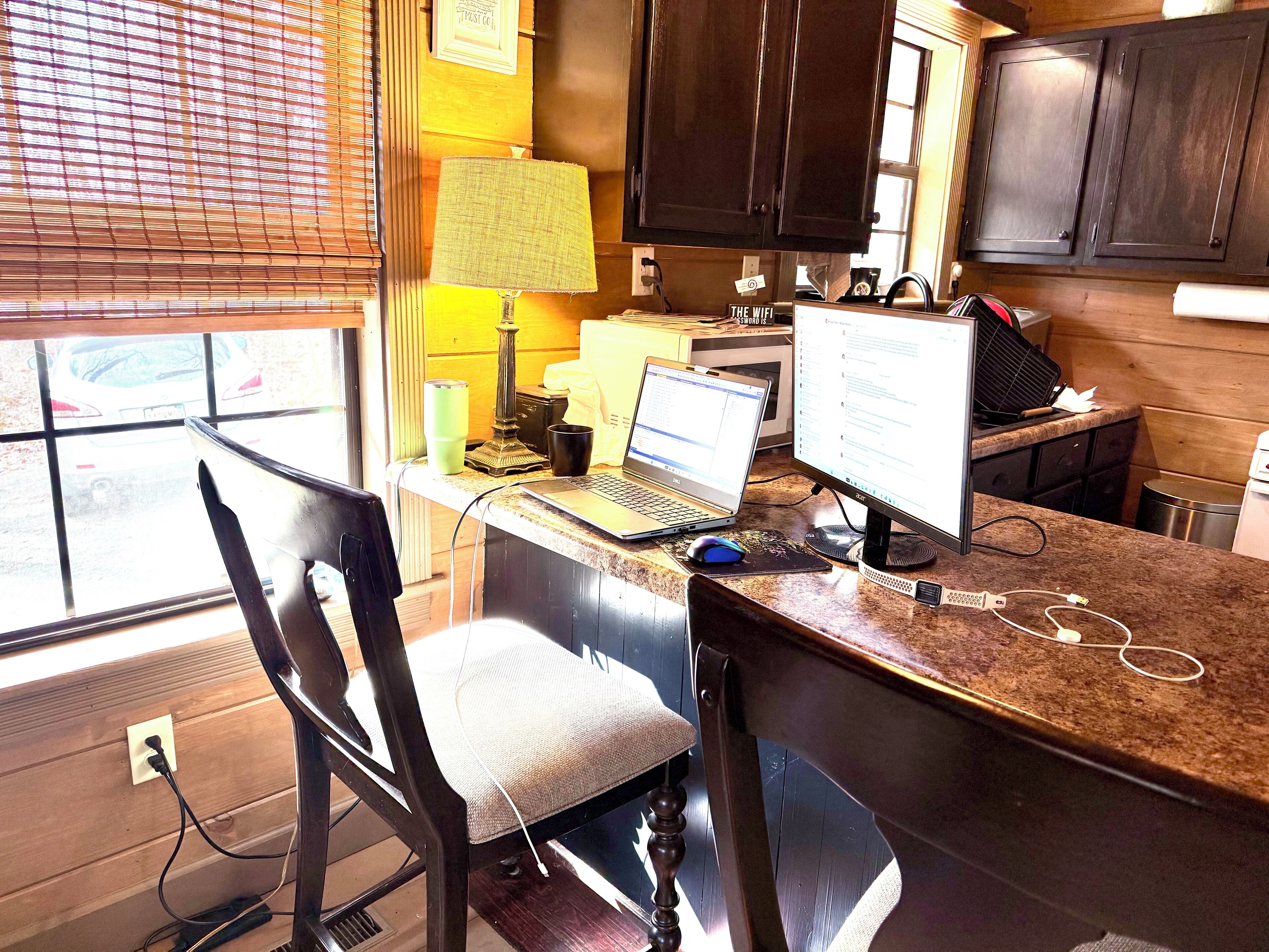 Una cocina con una computadora portátil y un computador de escritorio sobre la encimera, una silla y una ventana con persianas.