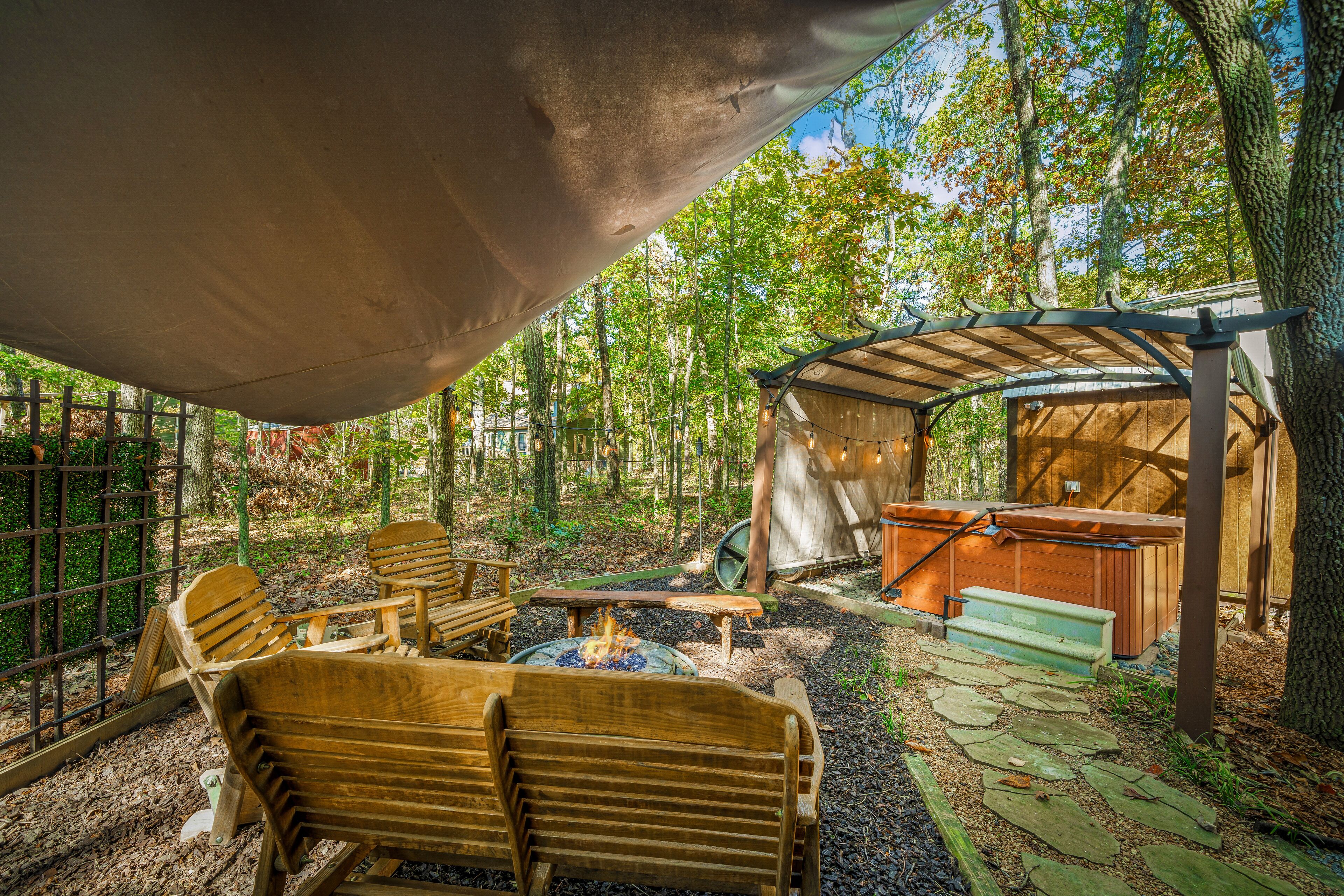 Un área de estar rústica al aire libre con muebles de madera y un brasero, rodeada de árboles y una estructura cubierta.