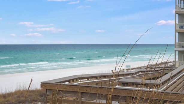 A beach view with a wooden boardwalk leading to a building with balconies.