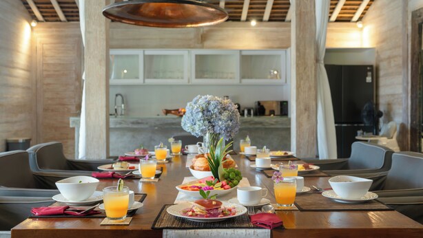 Desayuno preparado para dos personas, con una mesa dispuesta que incluye platos, vasos de jugo de naranja y un centro de mesa con flores.