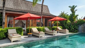 Un espace aménagé au bord de la piscine, avec des chaises longues et des parasols rouges, une piscine à l’eau claire et une végétation luxuriante.
