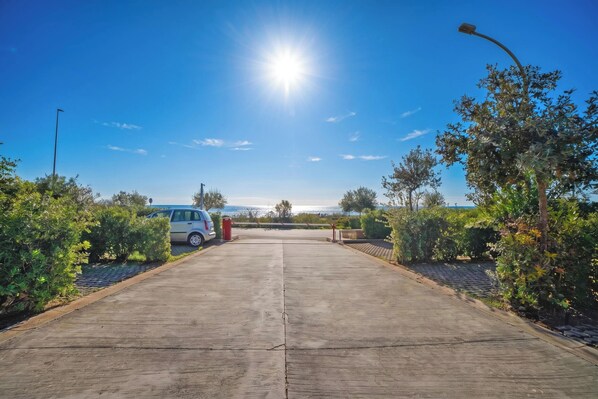 A paved pathway leading to a parking area with a car, surrounded by greenery and a clear sky.