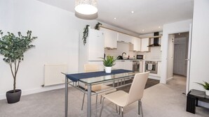 Une cuisine moderne avec des meubles blancs, un four à micro-ondes et une cuisinière. On y trouve un coin repas avec une table et des chaises en verre, ainsi qu’une plante en pot posée sur la table.