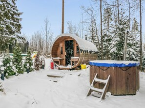 Une cabane en bois avec un toit enneigé, une terrasse en bois et un petit jacuzzi.
