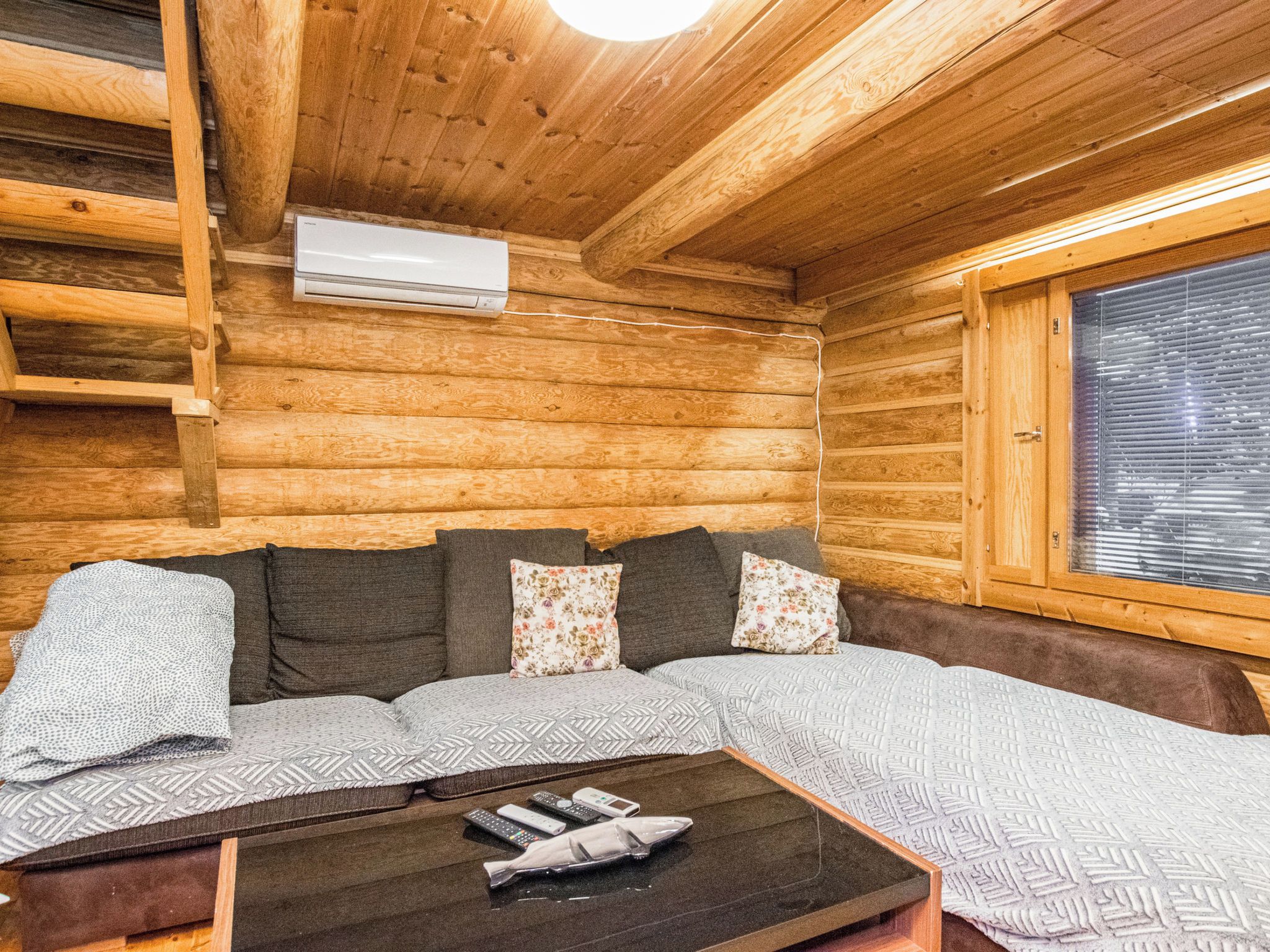 En stuga med träpanel på insidan, ett träbeklätt tak, en soffa med mönstrade kuddar, ett litet bord med fjärrkontroller och ett fönster med persienner.