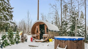 A wooden igloo structure with a bench and a small table inside, surrounded by snow-covered trees.