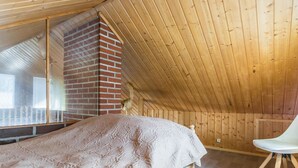 Makuuhuone, jossa on kalteva katto, tiilestä muurattu takka, kuviollinen päiväpeite ja valkoinen tuoli.