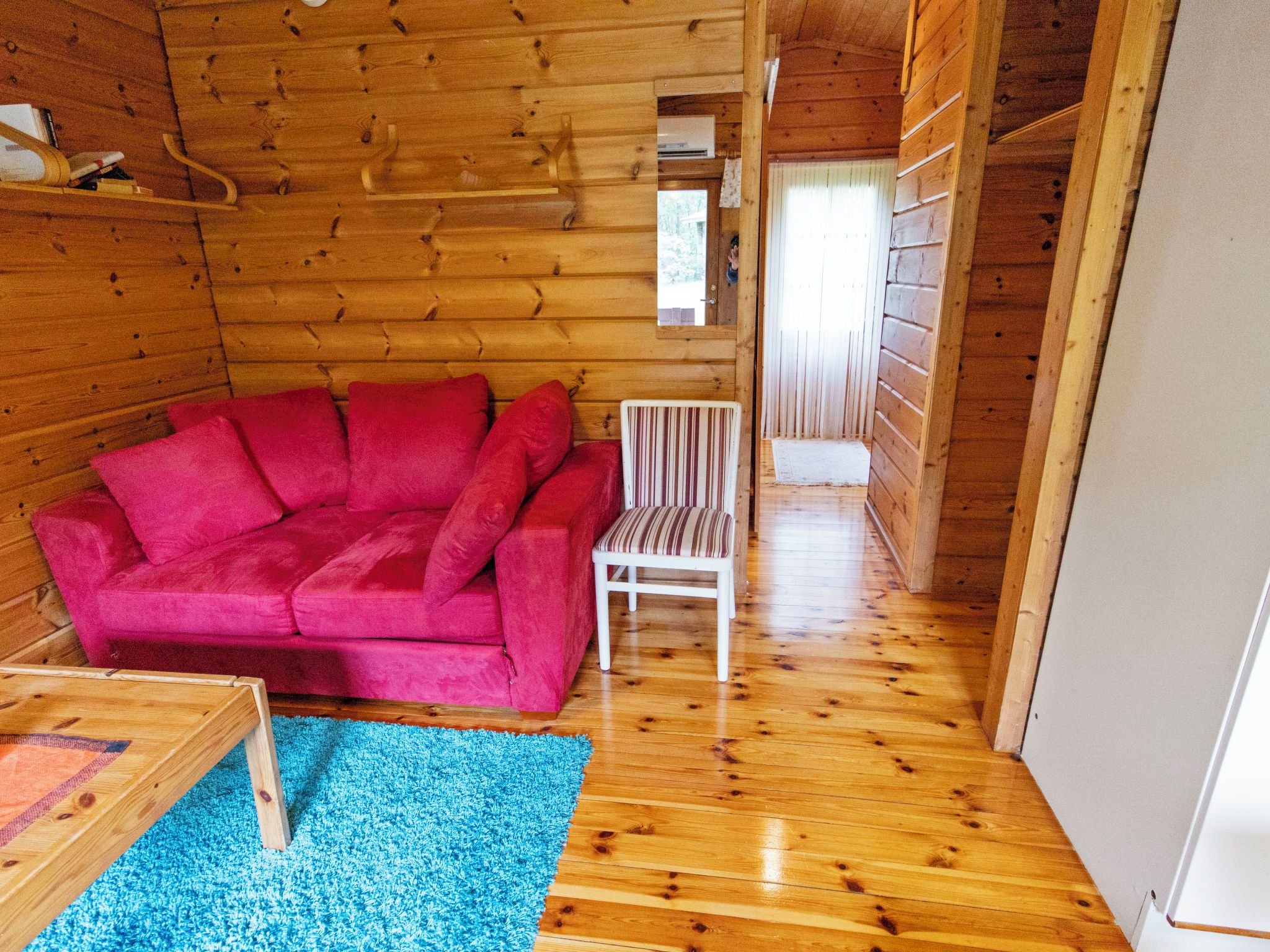 Interior de una cabaña de madera con un sofá rojo, una silla blanca y una alfombra azul.