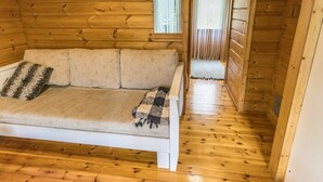 Interior de una cabaña de madera con un sofá, un ventanal pequeño y una puerta que da a otra habitación.