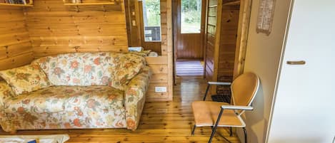 A wooden cabin interior with a sofa, a chair, a small table, and a refrigerator.
