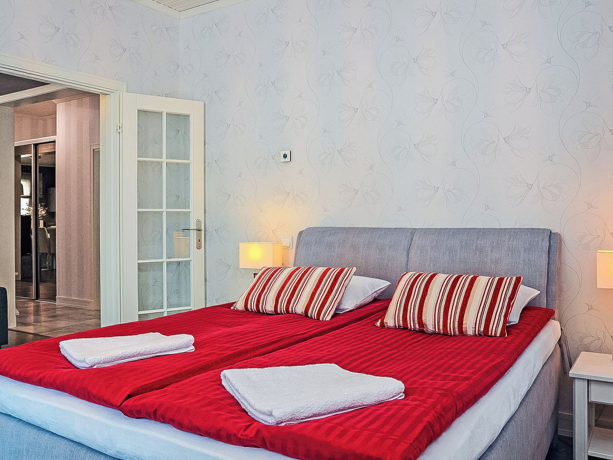 A double bed with red bedding and striped pillows, a bedside table with a lamp, and a door leading to a bathroom.