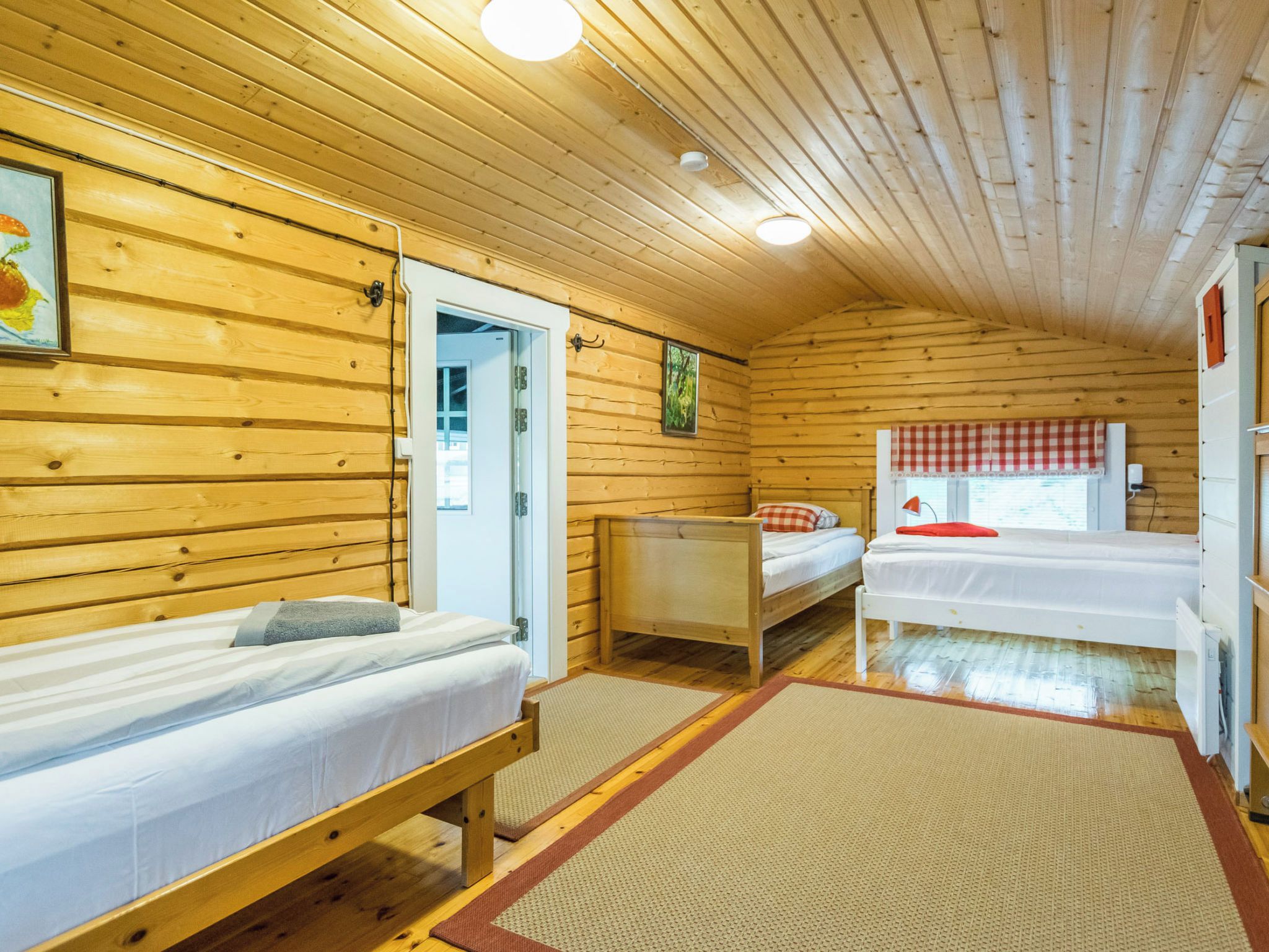 Una cabaña con paredes y pisos de madera, dos camas individuales, una mesita y una ventana con cortina a cuadros.