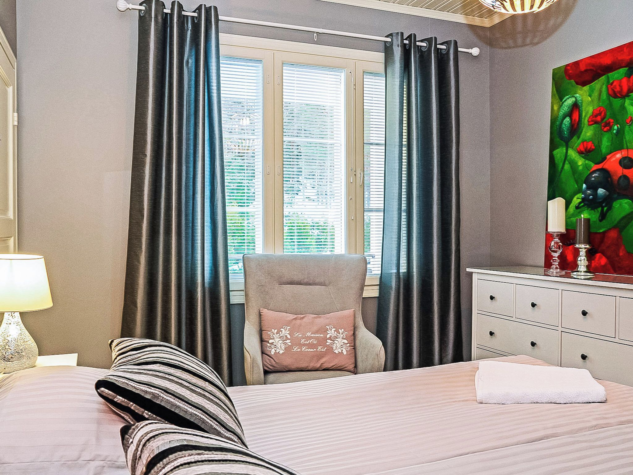 Ein Schlafzimmer mit Bett, Sessel, Kommode, einem Fenster mit Vorhängen und einem Bild an der Wand.