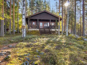 Eine Holzhütte, umgeben von einem Wald mit hohen Bäumen und einem mit Moos und Steinen bedeckten Boden.