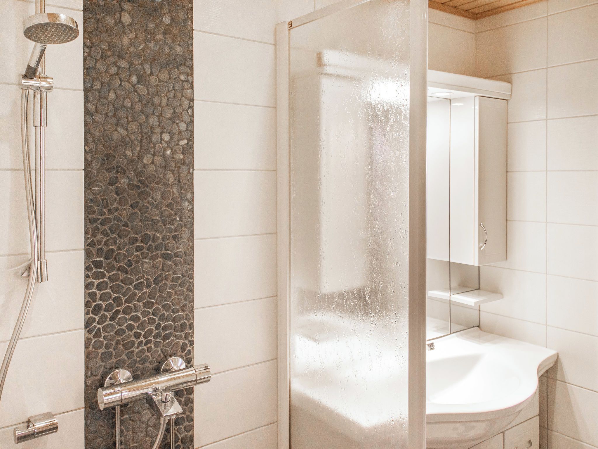 Une douche avec un mur en pierre, une porte en verre et une salle de bain carrelée de blanc.