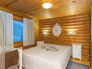 A cozy wooden cabin bedroom with a large bed, two bedside tables, a nightstand, a decorative mirror, and a window with curtains.