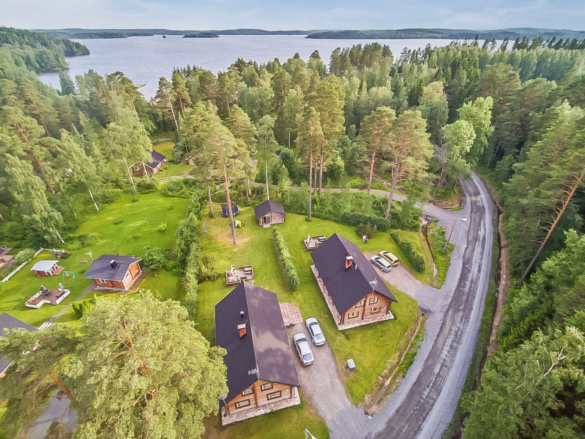 Un quartier résidentiel composé de plusieurs maisons, entouré d’une forêt dense, avec un lac en arrière-plan et une route sinueuse.